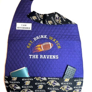 May include: Reversible purple quilted TV remote holder with the text "EAT, DRINK, WATCH THE RAVENS". Features a football graphic and pockets. The bottom and inside lining are black with the Baltimore Ravens logo.