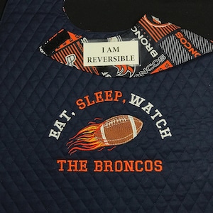 May include: Navy blue quilted bib with orange and white accents. The bib features embroidered text that reads "EAT, SLEEP, WATCH THE BRONCOS" with a football graphic. The bib is reversible and has a Broncos logo pattern.