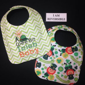 May include: A reversible baby bib with two designs. One side features a green and white chevron pattern with the embroidered text "A Wee Bit Irish Baby". The other side features a white background with a repeating pattern of green shamrocks, gold coins, and a cartoon leprechaun. The text "Leprechaun Luck!" is printed on the bib.