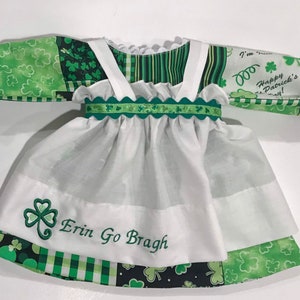 May include: A white dress with green shamrock print fabric and a white apron. The dress has a green ribbon with a scalloped edge and the words "Erin Go Bragh" embroidered on the apron. The sleeves are long and have a green shamrock print fabric. The dress is perfect for a St. Patrick's Day celebration.