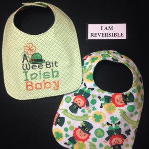 May include: A reversible baby bib with two sides. One side is green with white polka dots and embroidered text that says "A Wee Bit Irish Baby". The other side is white with a repeating pattern of green shamrocks, gold coins, and cartoon leprechauns with the text "Leprechaun Luck!"