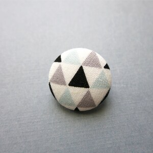 May include: A round button with a geometric pattern of black, gray, and light blue triangles.
