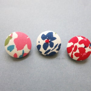 May include: Three round fabric buttons with floral patterns. The buttons are white with pink, blue, and red flowers.