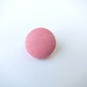 Puede incluir: Un botón redondo de color rosa claro cubierto de tela. El botón tiene una superficie texturizada y está sobre un fondo blanco. Este botón es adecuado para proyectos de costura y manualidades.