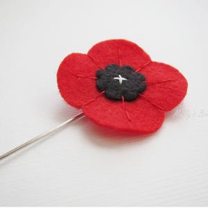 May include: A red felt poppy flower pin with a black center and a silver pin backing. The poppy has four petals and is stitched with red thread. The center has a white cross stitch.