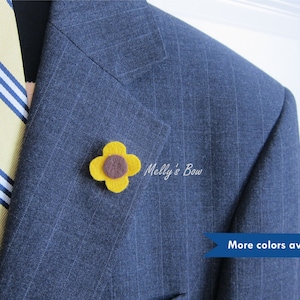 May include: A felt sunflower lapel pin in yellow and brown. The pin is on a blue suit jacket. "Melly's Bow" is written on the jacket. "More colors available" is written below the pin.