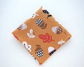 May include: Folded square of fabric with a repeating pattern of stylized acorns, leaves, and flowers in white, brown, red, and black on a golden-orange background. The fabric is likely cotton or a similar material.
