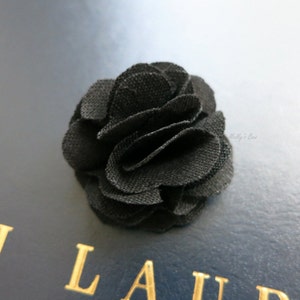 Könnte beinhalten: Ein schwarzer Stoffblumen-Anstecker. Die Blume besteht aus mehreren Stoffschichten und hat eine gekräuselte Optik.