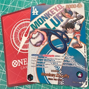 May include: A trading card featuring the character Monkey D. Luffy from the "One Piece" series, holding a baseball and bat. The card has a blue and white background with the text "MONKEY.D.LUFFY" and "LA" logo. The card is red on the back.