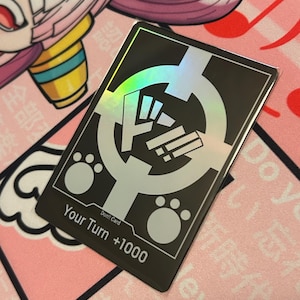 May include: A black and silver card with a holographic finish. The card has a white paw print on each side and the text "Don!! Card Your Turn +1000" in white.
