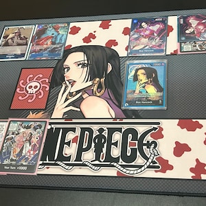 Puede incluir: Un tapete de juego de cartas coleccionables de One Piece con una ilustraci&oacute;n en blanco y negro del logotipo de One Piece sobre un fondo rojo y blanco. El tapete tambi&eacute;n presenta un personaje de la serie de anime, Boa Hancock, con un vestido morado con un s&iacute;mbolo de calavera y huesos cruzados en el fondo.