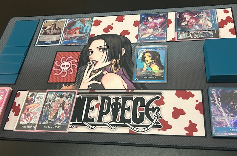 Puede incluir: Un tapete de juego de cartas coleccionables de One Piece con una ilustraci&oacute;n en blanco y negro del logotipo de One Piece sobre un fondo rojo y blanco. El tapete tambi&eacute;n presenta un personaje de la serie de anime, Boa Hancock, con un vestido morado con un s&iacute;mbolo de calavera y huesos cruzados en el fondo.