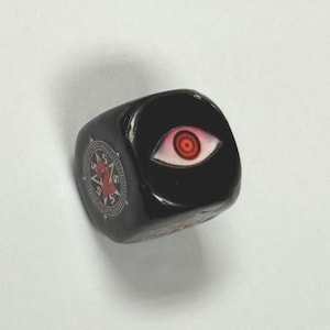 May include: A black, six-sided die featuring a red and white eye design on one face and a red and white emblem on another. The die is glossy and appears to be made of a hard material.