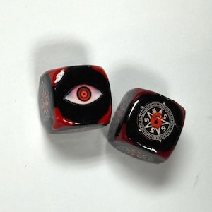 Może przedstawiać: Dwie czarne kostki z czerwonymi akcentami. Jedna kostka przedstawia białe oko z czerwoną tęczówką, druga wzór róży wiatrów. Kostki są prawdopodobnie używane do gier planszowych lub RPG.