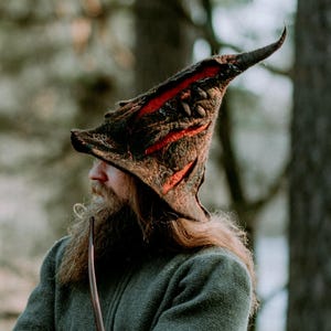 Könnte beinhalten: Ein spitzer, strukturierter brauner Hut mit leuchtend orangefarbenen Innenakzenten, getragen zu einem dunkelolivgrünen Reißverschluss-Pullover, dunkler Hose und einer braunen Ledertasche mit geprägten Mustern an der Seite.