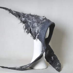 May include: A gray felt hood with a long, pointed tail and white curly accents. The hood is displayed on a white mannequin head. The felt has a textured appearance, and the design is reminiscent of a medieval or fantasy style.