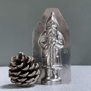 Puede incluir: Un molde de metal plateado para hacer figuras de Papá Noel de chocolate. El molde tiene la forma de un Papá Noel de pie con una bolsa de regalos y una barba. El molde tiene el número 120 impreso.