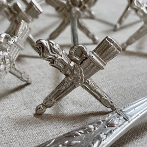 May include: Silver-colored metal objects with a crossed torch design. The torches have detailed textures and are likely decorative items. The image shows multiple pieces, suggesting a set or collection.