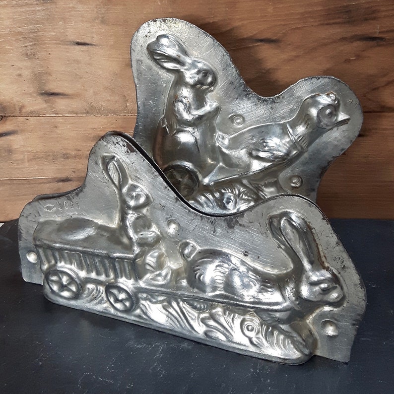 May include: Two vintage metal molds for making chocolate bunnies and a chick. The molds are silver in color and have intricate details. One mold features a bunny pulling a wagon with a chick inside. The other mold features a bunny and a chick side by side.