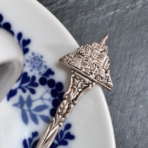 May include: A silver-colored souvenir spoon with a detailed miniature cityscape design. The spoon rests on a white plate with blue floral patterns. The spoon's handle has an ornate, decorative design.
