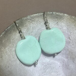 May include: A pair of dangle earrings featuring smooth, light mint green ceramic beads with a subtle crackle glaze finish. Each bead is suspended from a silver-toned metal hook wire, accented with small clear glass spacer beads at the top and bottom.