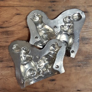 May include: Two vintage metal molds for making chocolate bunnies and birds. The molds are made of silver-colored metal and have a detailed design.