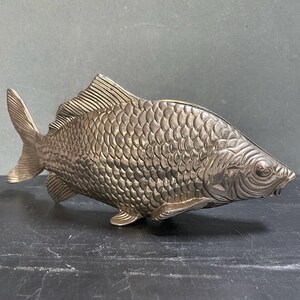 May include: A silver-colored fish sculpture with detailed scales, fins, and a tail. The fish is positioned horizontally, showcasing its textured surface and design. The sculpture is likely a decorative item.