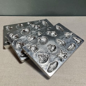 May include: Three silver-colored metal molds with various seashell shapes. The molds are square and have a raised design of different types of shells, starfish, and other sea creatures. The molds are stacked on top of each other.