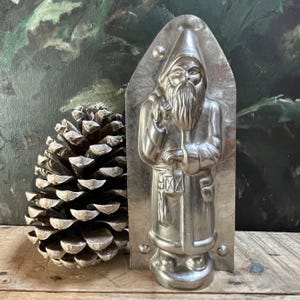 Könnte beinhalten: Eine silberfarbene Metallform des Weihnachtsmanns mit langem Bart, spitzer Mütze, einem Sack über der Schulter und einer Laterne in der Hand. Die Form hat eine abgerundete Oberseite, einen flachen Boden und kleine kreisförmige Vertiefungen.