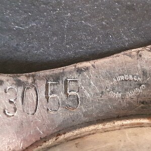 May include: Close-up of a silver-colored metal object with the number "3055" stamped on it. The object appears to be part of a larger piece of metal.