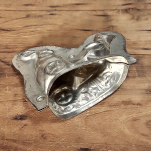 May include: A vintage metal mold for making chocolate candy. The mold is shaped like a person riding a bicycle. The mold is silver in color and has a worn, antique look.