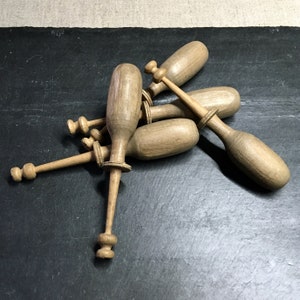 Op de afbeelding: Een groep van vijf houten spoelen voor kantklossen, gerangschikt op een donkergrijs oppervlak. De spoelen zijn allemaal van verschillende grootte en vorm en hebben een natuurlijke houten afwerking.