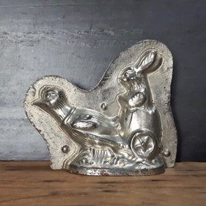 May include: A vintage metal mold for making chocolate bunnies and chicks. The mold is silver in color and features a detailed design of a rabbit pulling a cart with a chick.