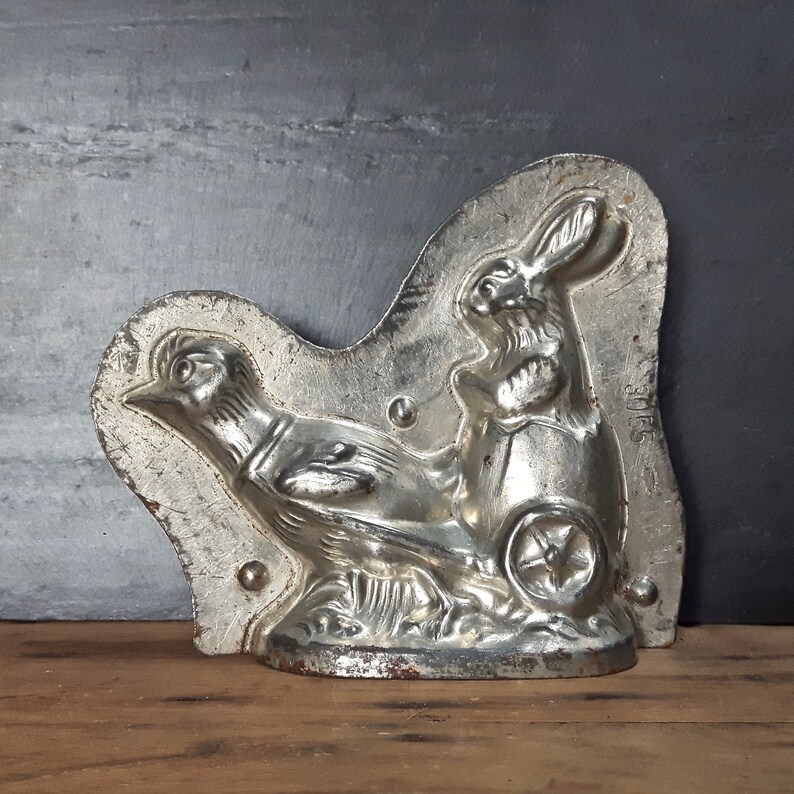 May include: A vintage metal mold for making chocolate bunnies and chicks. The mold is silver in color and features a detailed design of a rabbit pulling a cart with a chick.