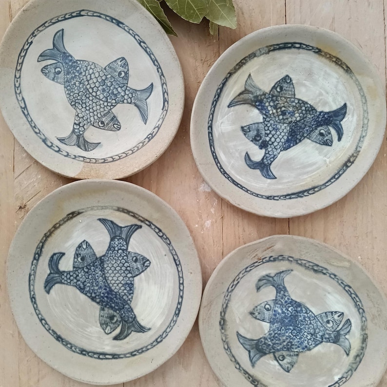 May include: Four round ceramic plates with a cream-colored glaze and a hand-painted blue design. Each plate features a central motif of three interlocking fish with detailed scales, surrounded by a decorative blue border along the rim.