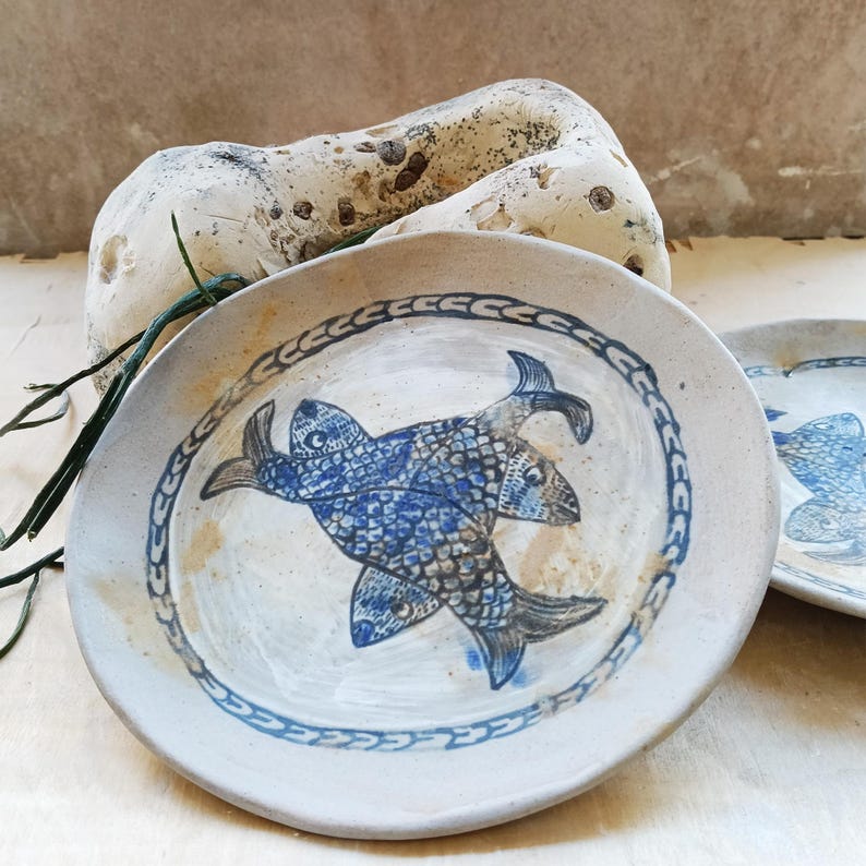 May include: A round, off-white ceramic plate featuring a hand-painted blue design of two overlapping fish in the center. The plate has a decorative blue border pattern around the rim and a slightly uneven, organic edge, showcasing a textured finish.