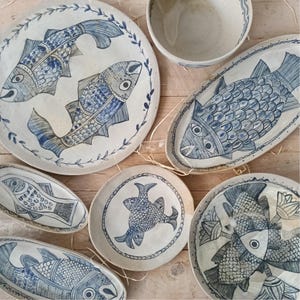 May include: A collection of off-white ceramic plates and bowls featuring hand-painted blue fish motifs. The set includes round and oval dishes decorated with detailed fish scales, fins, and leafy borders, showcasing a consistent blue and white color palette.