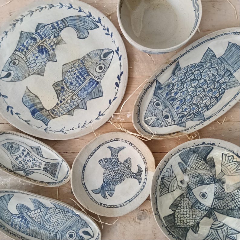 May include: A collection of off-white ceramic plates and bowls featuring hand-painted blue fish motifs. The set includes round and oval dishes decorated with detailed fish scales, fins, and leafy borders, showcasing a consistent blue and white color palette.