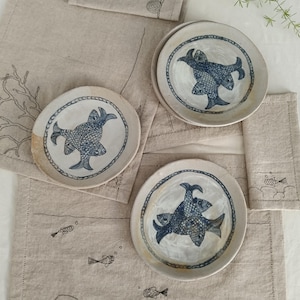 May include: Three round ceramic plates with a cream-colored glaze and dark blue hand-painted fish illustrations, arranged on a beige linen runner featuring embroidered fish and coral motifs, creating a cohesive set of nautical-themed tableware.