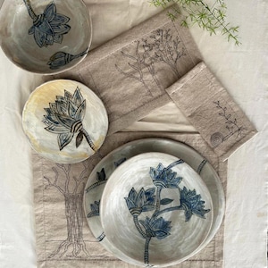 Op de afbeelding: Een set keramieken servies bestaande uit een kom en twee borden met handgeschilderde blauwe lotusbloemen, gerangschikt op beige linnen placemats en een servet met zwarte lijntekeningen van bomen en een slak.