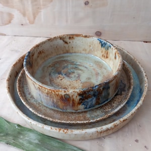 Può includere: Un set di tre ciotole in ceramica in varie tonalità di marrone, blu e bianco. Le ciotole sono annidate, con la ciotola più grande in basso e la più piccola in alto.