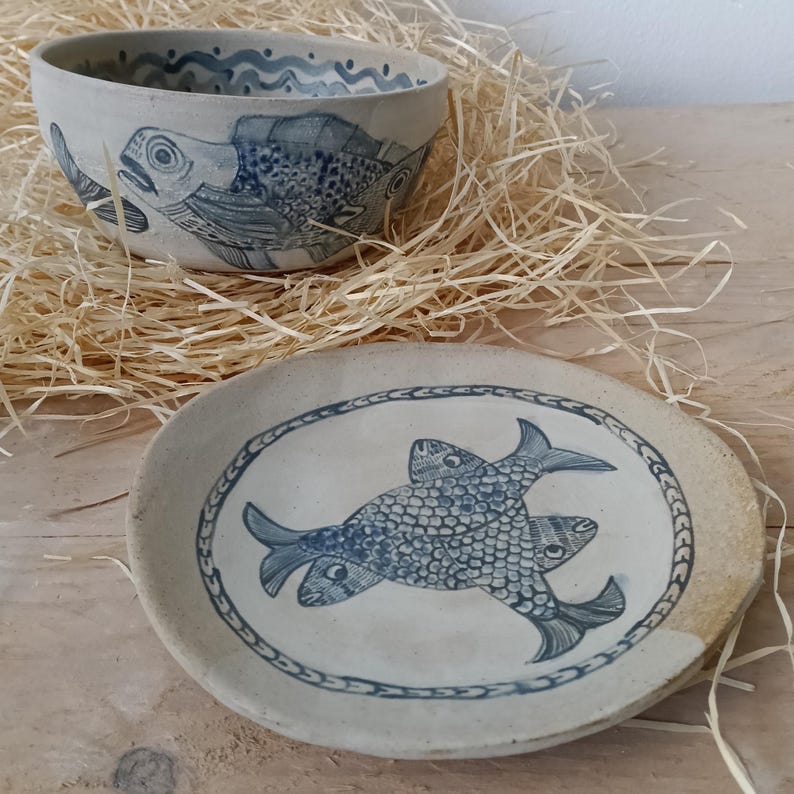 May include: A ceramic bowl and a matching plate featuring hand-painted blue fish illustrations on a light beige background. The bowl has a wavy line pattern along the inner rim, while the plate displays two overlapping fish within a circular border.