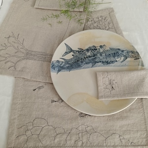 Könnte beinhalten: Ein runder Keramikteller mit einer blauen Illustration eines Fischschwarms, platziert auf hellbeigen Leinen-Tischsets. Die Tischsets sind mit schwarzen Strichzeichnungen von Bäumen, Felsen und kleinen Fischen sowie gesäumten Kanten verziert.