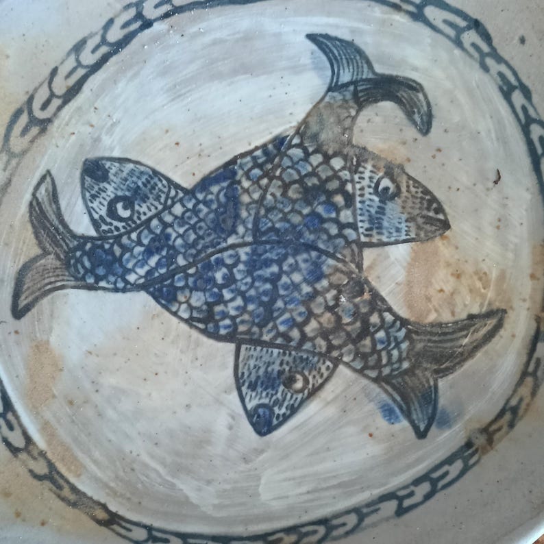 May include: A round ceramic plate featuring a hand-painted design of three interlocking fish in shades of blue and grey. The fish have detailed scale patterns and are arranged in a circular formation. The plate has a cream-colored base with a decorative border.