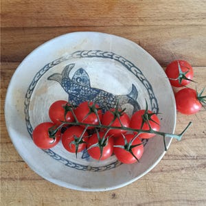 May include: A round, off-white ceramic plate featuring a dark blue hand-drawn fish illustration in the center and a decorative border pattern. A vine of eight bright red cherry tomatoes with green stems rests across the surface of the plate.