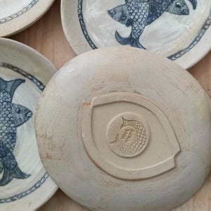 May include: A collection of round ceramic plates featuring blue hand-painted fish illustrations. The underside of one plate displays a central embossed stamp with a fish motif and the text TINA POUPE, surrounded by a raised almond-shaped border.
