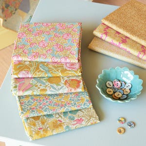 May include: A stack of floral fabric squares in pink, blue, and yellow, with a small turquoise bowl of buttons. Additional fabric squares with a gold and pink pattern are stacked nearby. The fabrics are arranged on a light blue table.
