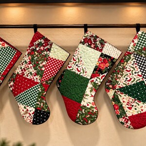 May include: Four quilted Christmas stockings hanging in a row, each featuring a patchwork design of red, green, white, and black fabrics. The patterns include gingham, floral prints, and holiday motifs, all finished with black binding and hanging loops.
