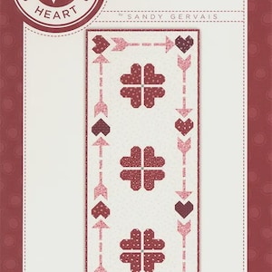 May include: A quilt pattern titled "Cupid's Presence" by Sandy Gervais. The design features a central panel with four-leaf clover shapes and arrows, hearts, and dotted backgrounds in shades of red and pink.