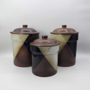 May include: A set of three ceramic canisters with lids, featuring a geometric pattern of cream, dark brown, and black triangular segments. Each cylindrical container has a smooth, glazed finish and a matching brown lid with a small knob handle.